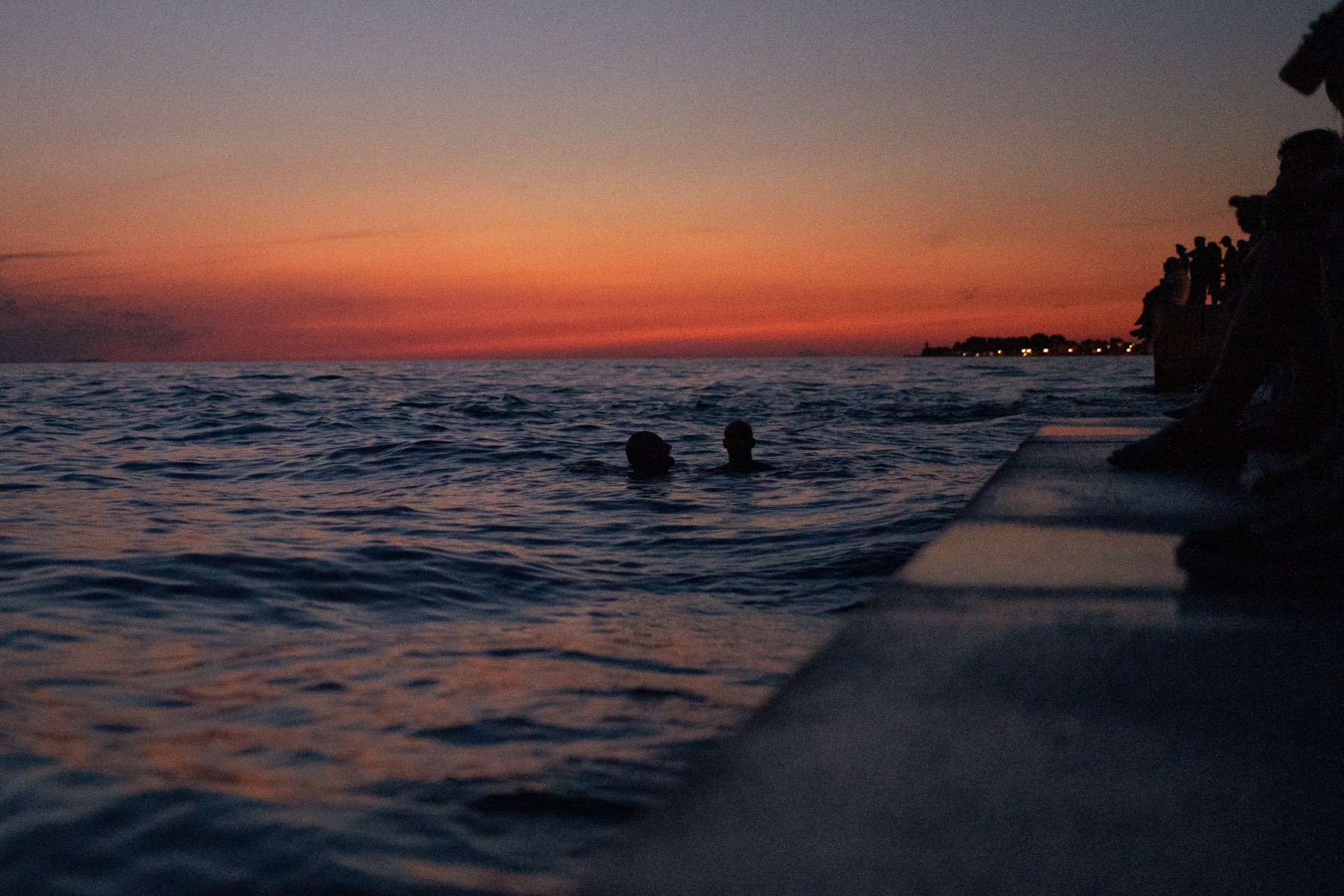 Two swimmers silhouetted against an orange sunset over the sea.